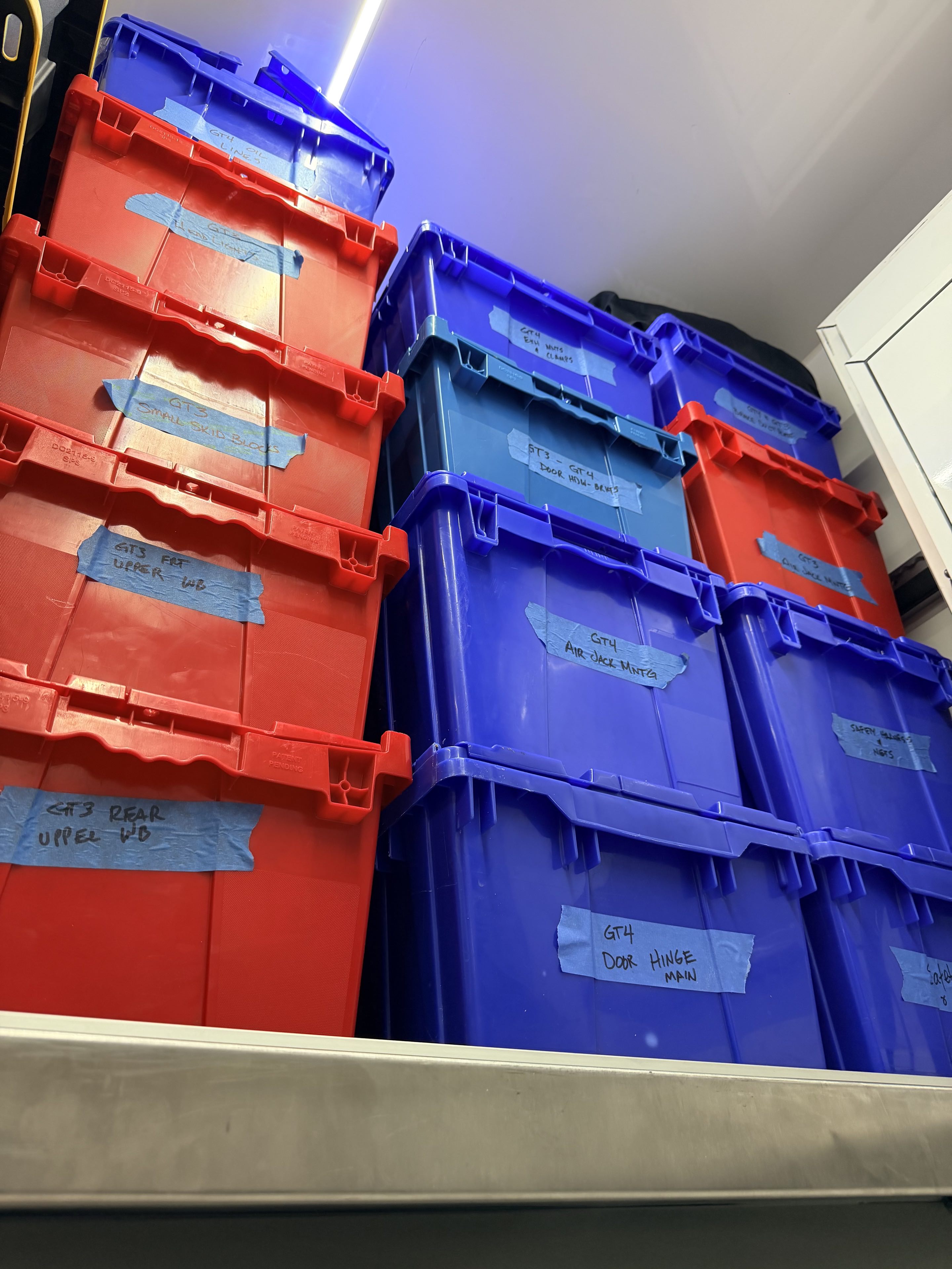 Blue and red plastic bins stacked to the ceiling with tape labels indicating various race car parts